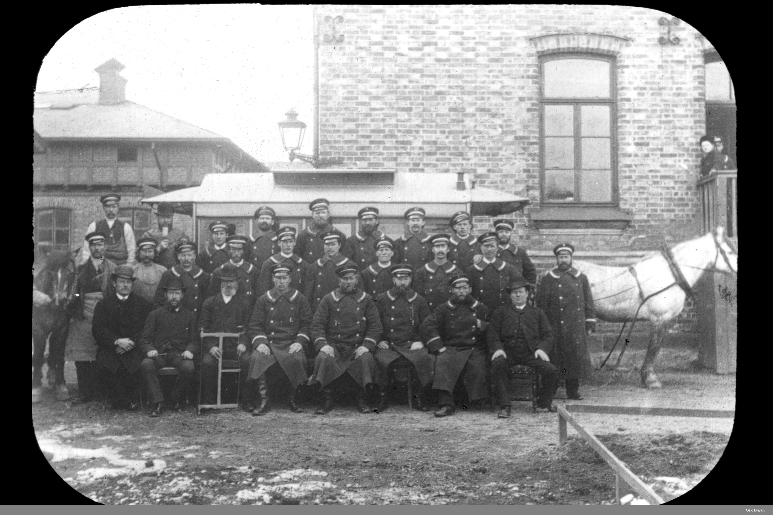 Sort-hvitt av menn i uniform foran hest og hestesporvogn