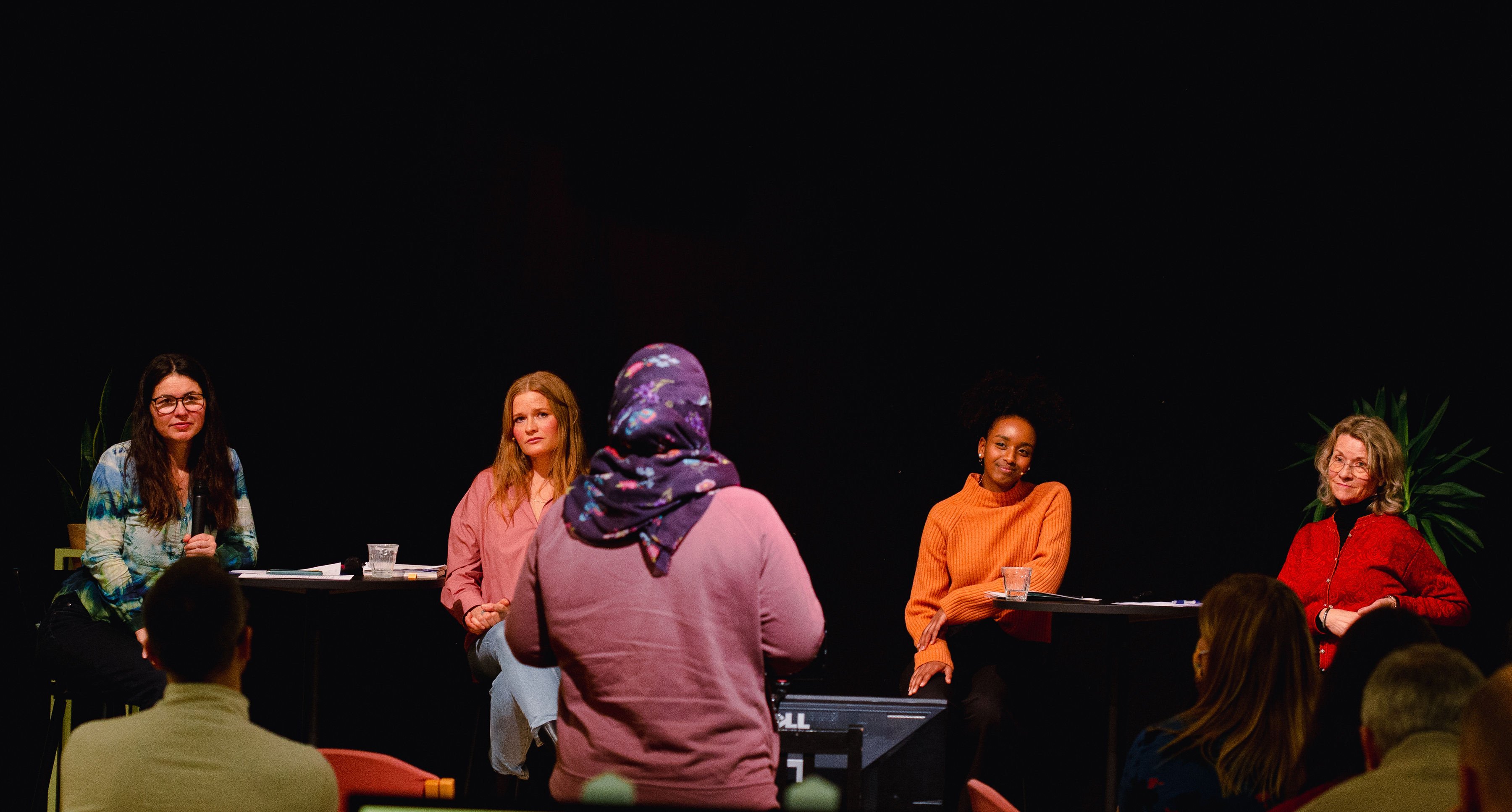 Fire kvinner sitter på hver sin stol i en panelsamtale på en liten scene med ansiktene mot kamera. En kvinne med hijab står vendt mot dem.