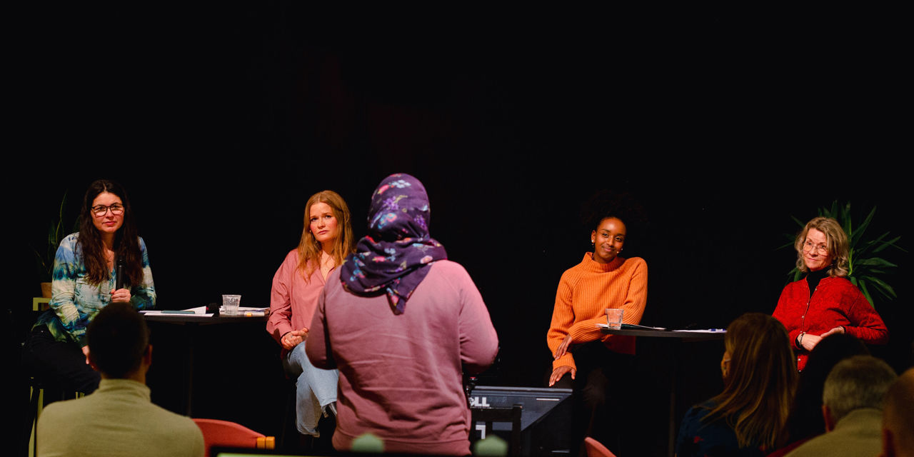 Fire kvinner sitter på hver sin stol i en panelsamtale på en liten scene med ansiktene mot kamera. En kvinne med hijab står vendt mot dem.