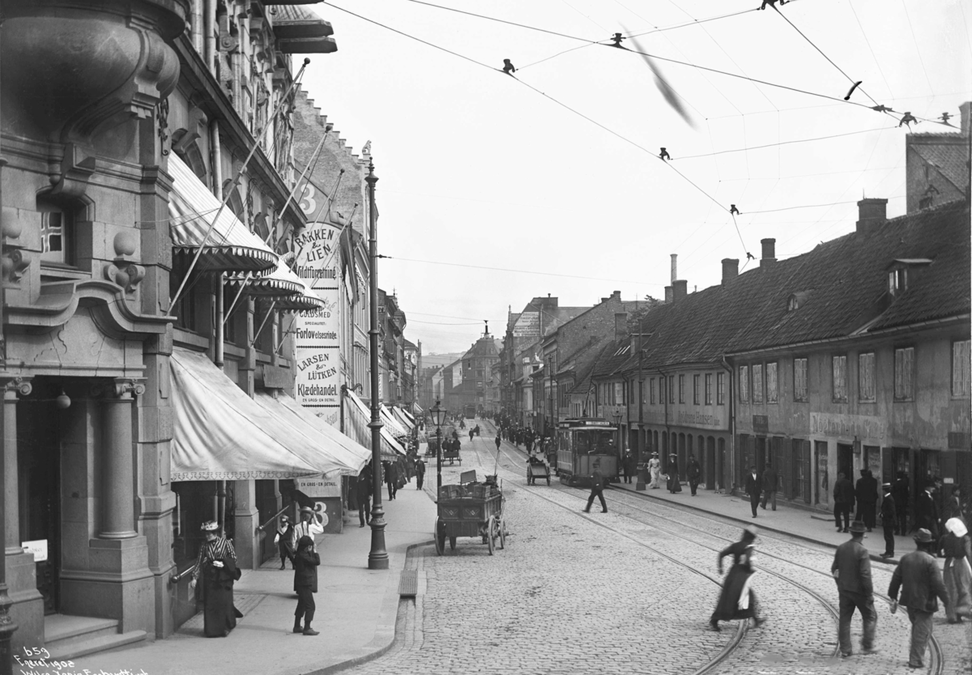 Sort-hvitt-bilde fra 1902 av Storgata i Oslo med mennesker, trikkeskinner og hestevogner.