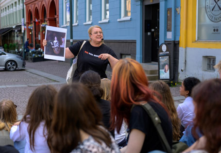 Kvinne med Oslo Museum-tskjorte står i gate foran ungdom og holder opp et gammelt portrettbilde