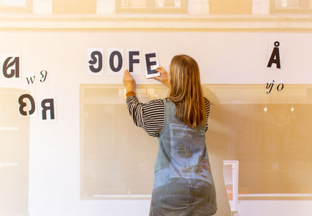 Bygningsfasade projisert på en vegg i museet. Ung kvinne lager ord med magnetiske bokstaver som ikke eksisterer i norsk språk i dag.