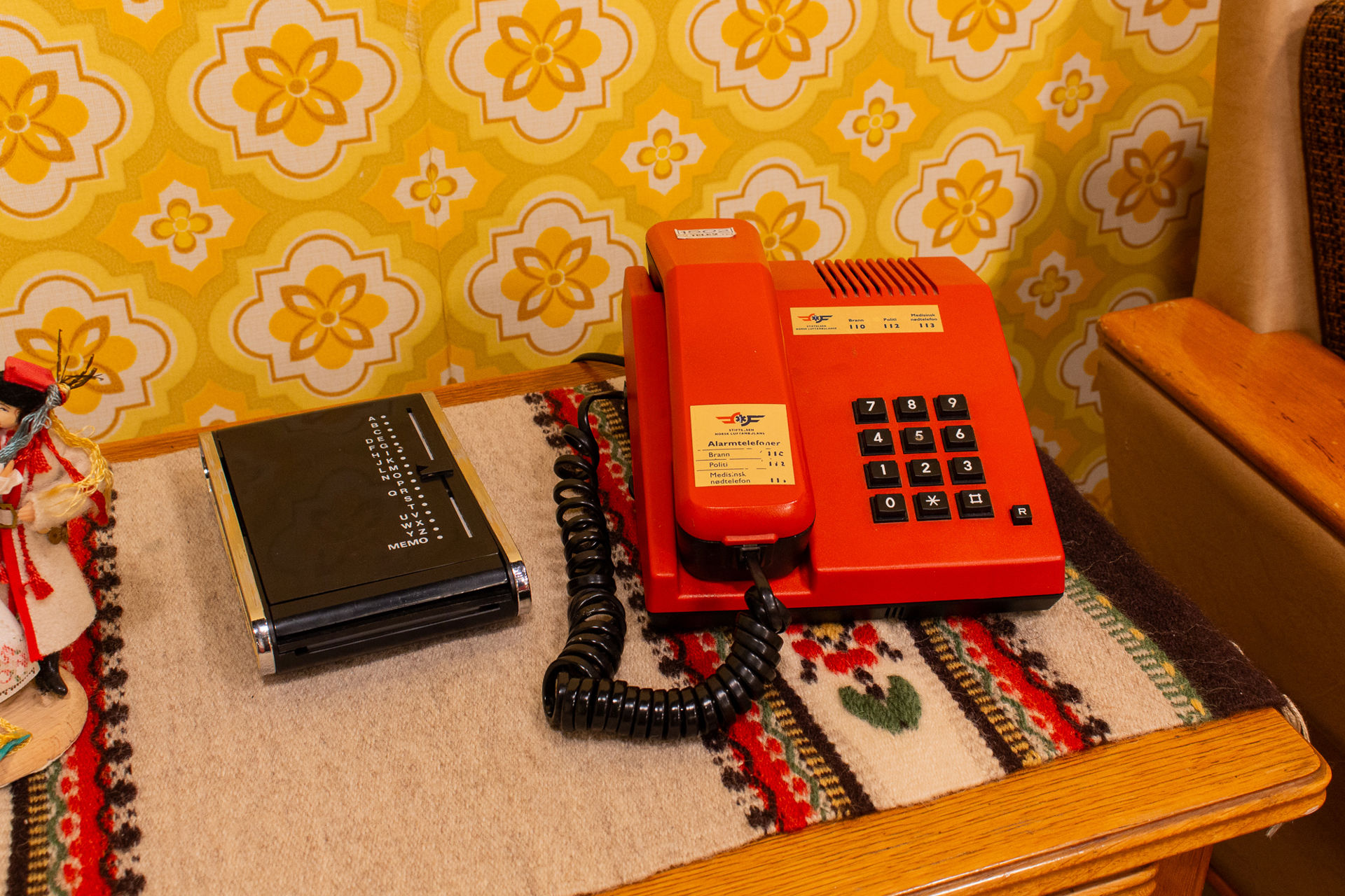 Rød fasttelefon og telefonbok på brodert duk foran et gult blomstertapet