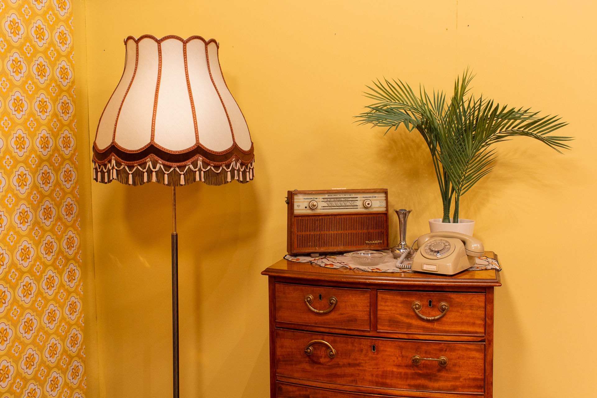 Hvit, retro lampe ved siden av en trekommode med telefon med tallskive, en grønn plante og en gammel radio