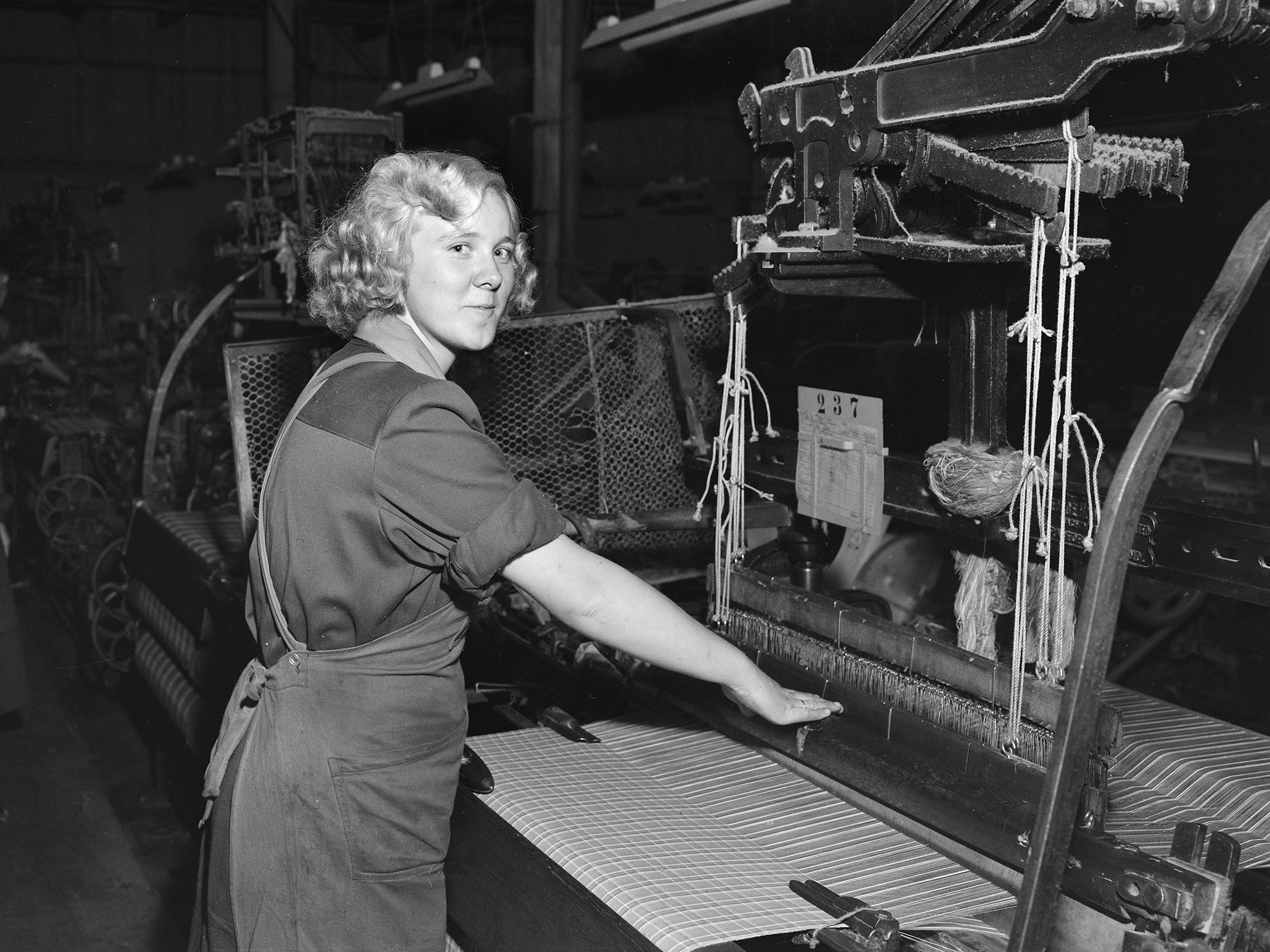 Ung kvinne betjener vevmaskin. Sort-hvitt bilde.