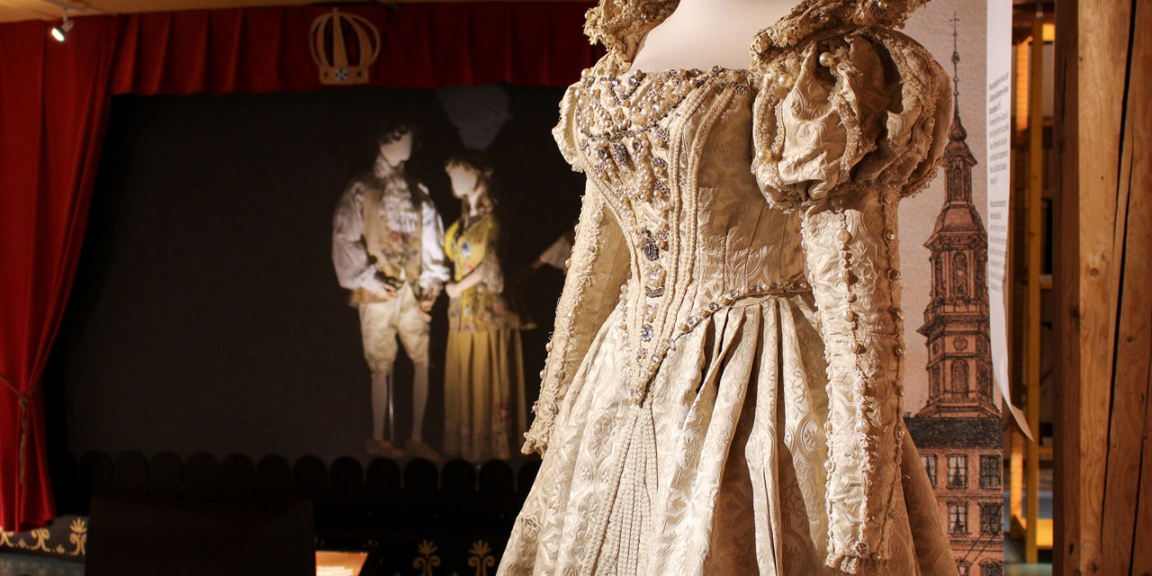En flott kjole er hovedmotivet. I bakgrunnen to utstillingsdukker med teaterbekledning fra gamle dager.