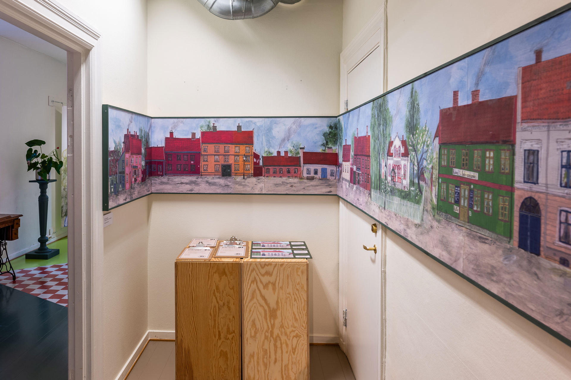 Hvitt lite rom med to trekasser med oppgaveark i midten. En stor avlang illustrasjon av gamle fargerike trehus går over alle tre veggene.