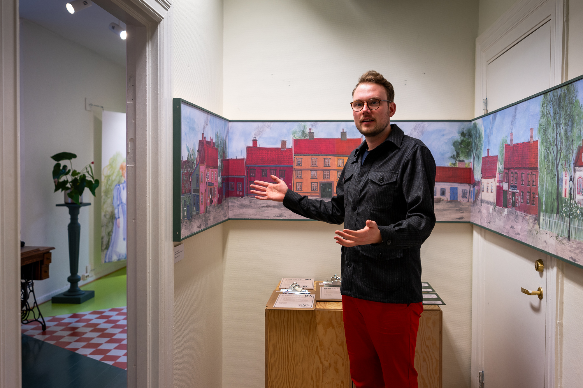 Mann snakker og står med armene ut foran en stor illustrasjon og fargerike trehus.