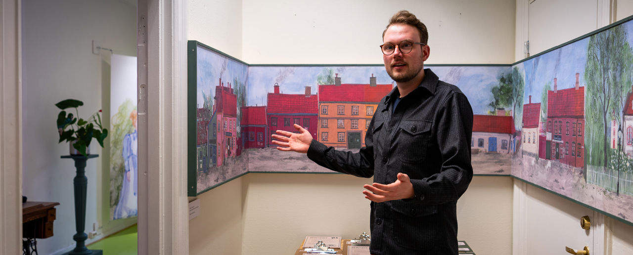 Mann snakker og står med armene ut foran en stor illustrasjon og fargerike trehus.