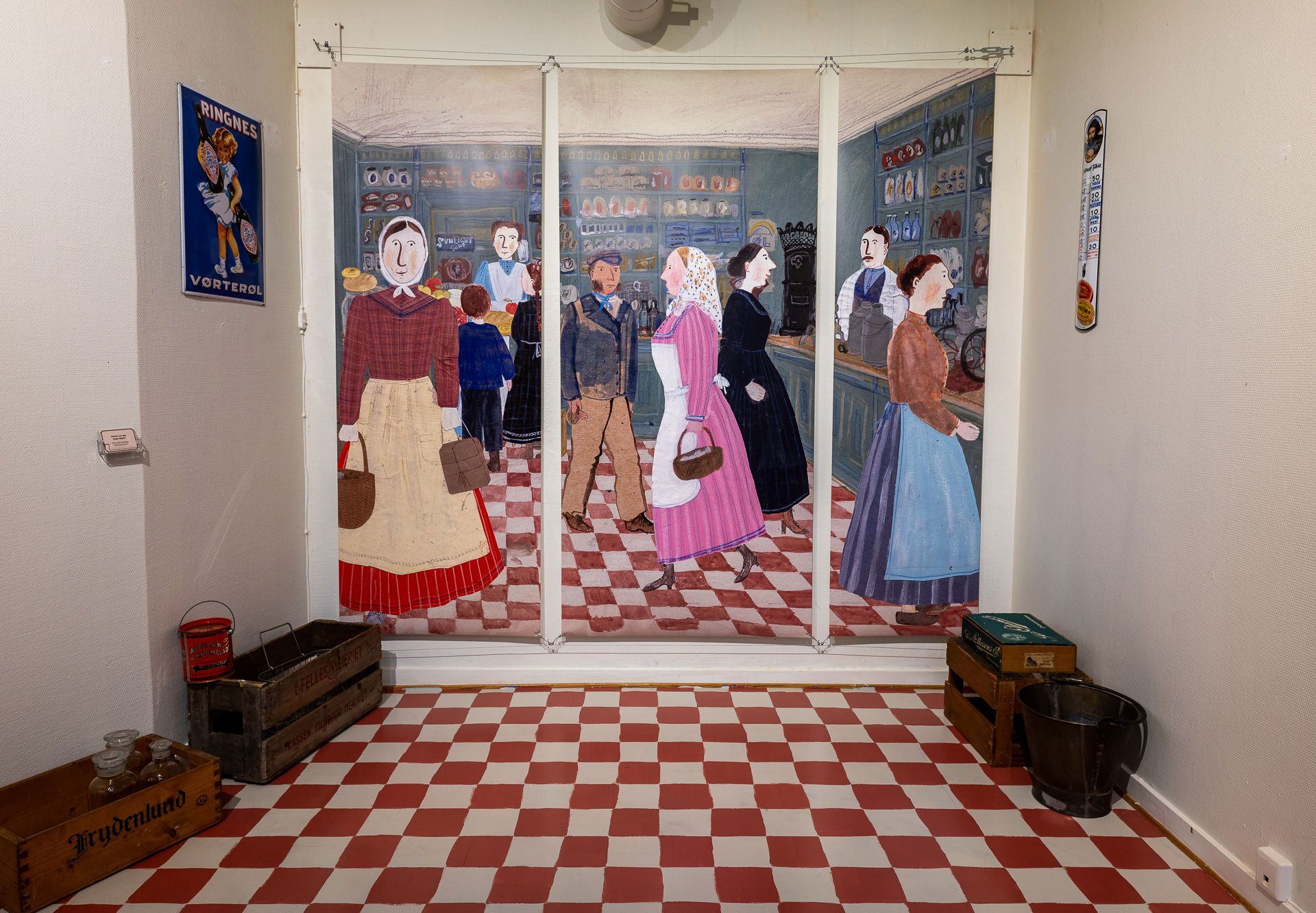 Rom med hvite vegger og rødrutete gulv, gamle trekasser på gulvet, stor bannerillustrasjon midt på veggen av personer ikledd gammeldagse klær i en butikk.