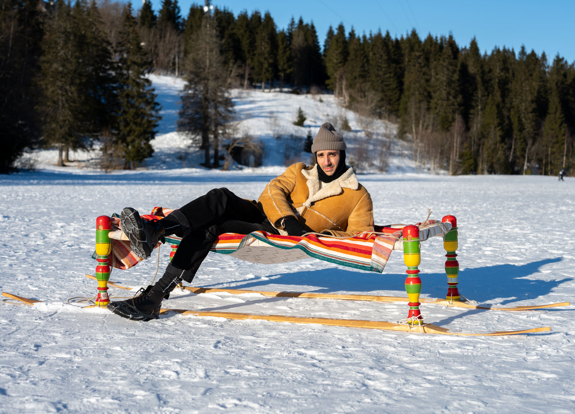 Mann i saueskinnsjakke ser mot kamera mens han ligger sidelengs på en dagseng med fargerike bein og treski montert på beina i snøen