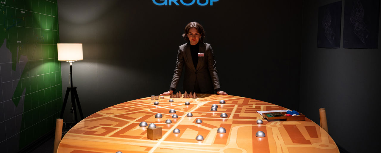 Dame i dress i et mørkt rom lener seg på et stort spillbord mens hun er i kamera. Thorn Group står i store blå bokstaver på veggen bak.