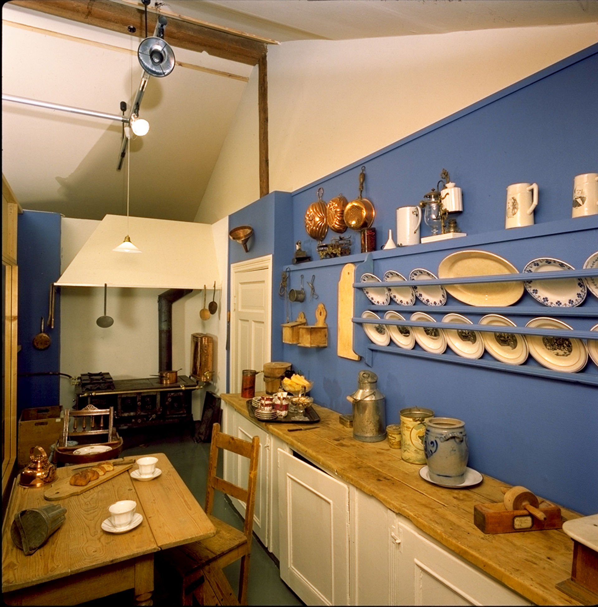 Gammelt kjøkken med blå vegg og naturlig trebord. Asjetter og kjeler henger på den blå veggen.