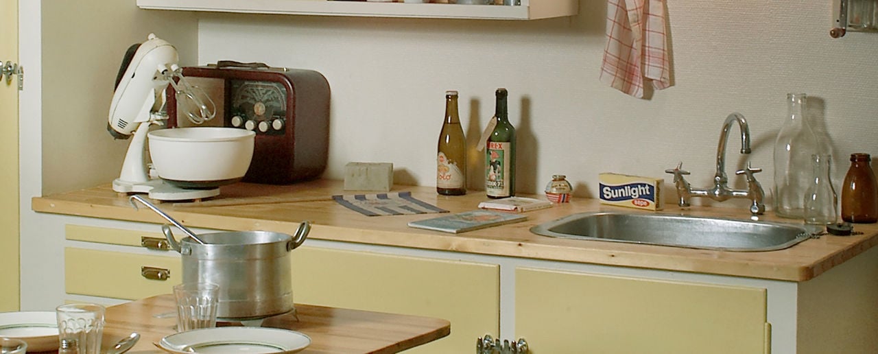 Kjøkken fra 50-tallet, gule skapdører, overskapet har skyvedører. En kjele og tallerken står på et bord i forkant.