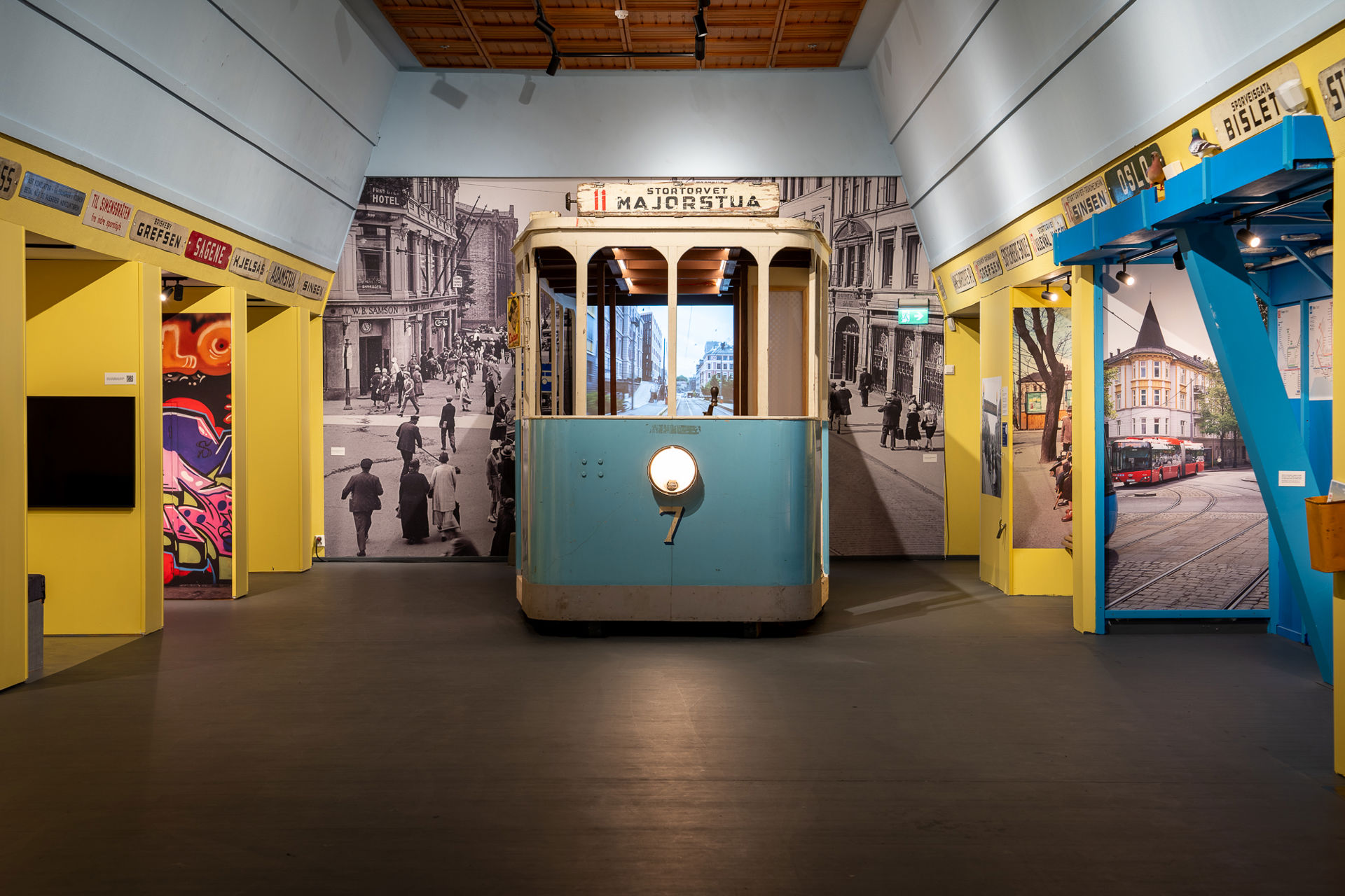 En gammel blå trikkemodell plassert midt i rommet med et sort-hvitt-bybilde bak. Gule vegger rundt med skilt med ulike stedsnavn i Oslo.