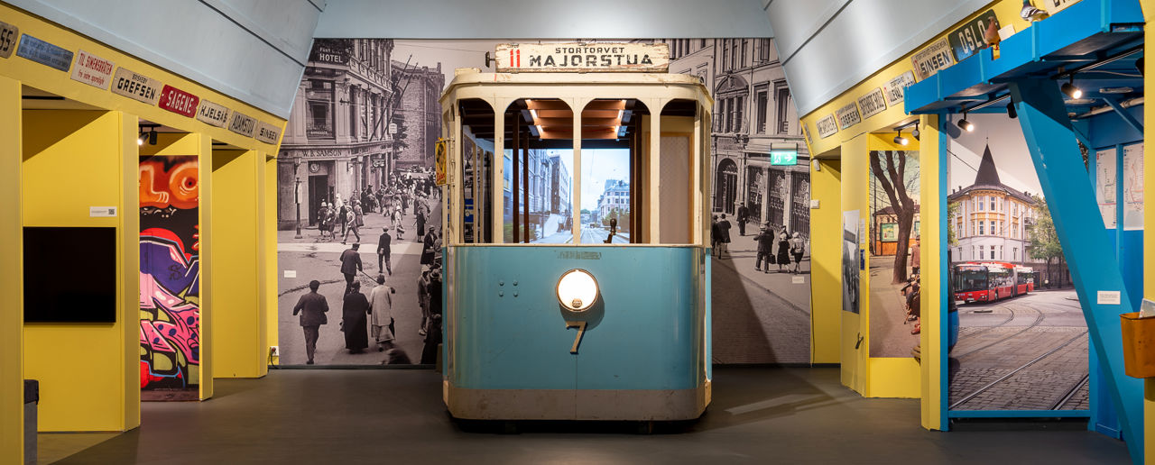 En gammel blå trikkemodell plassert midt i rommet med et sort-hvitt-bybilde bak. Gule vegger rundt med skilt med ulike stedsnavn i Oslo.