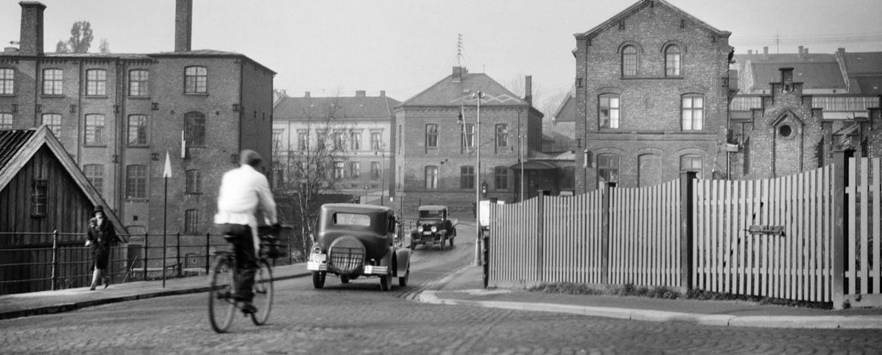 Sort-hvitt-bilde av mann som sykler etter en gammel bil over en bru. Fabrikkbygninger på hver side av brua.