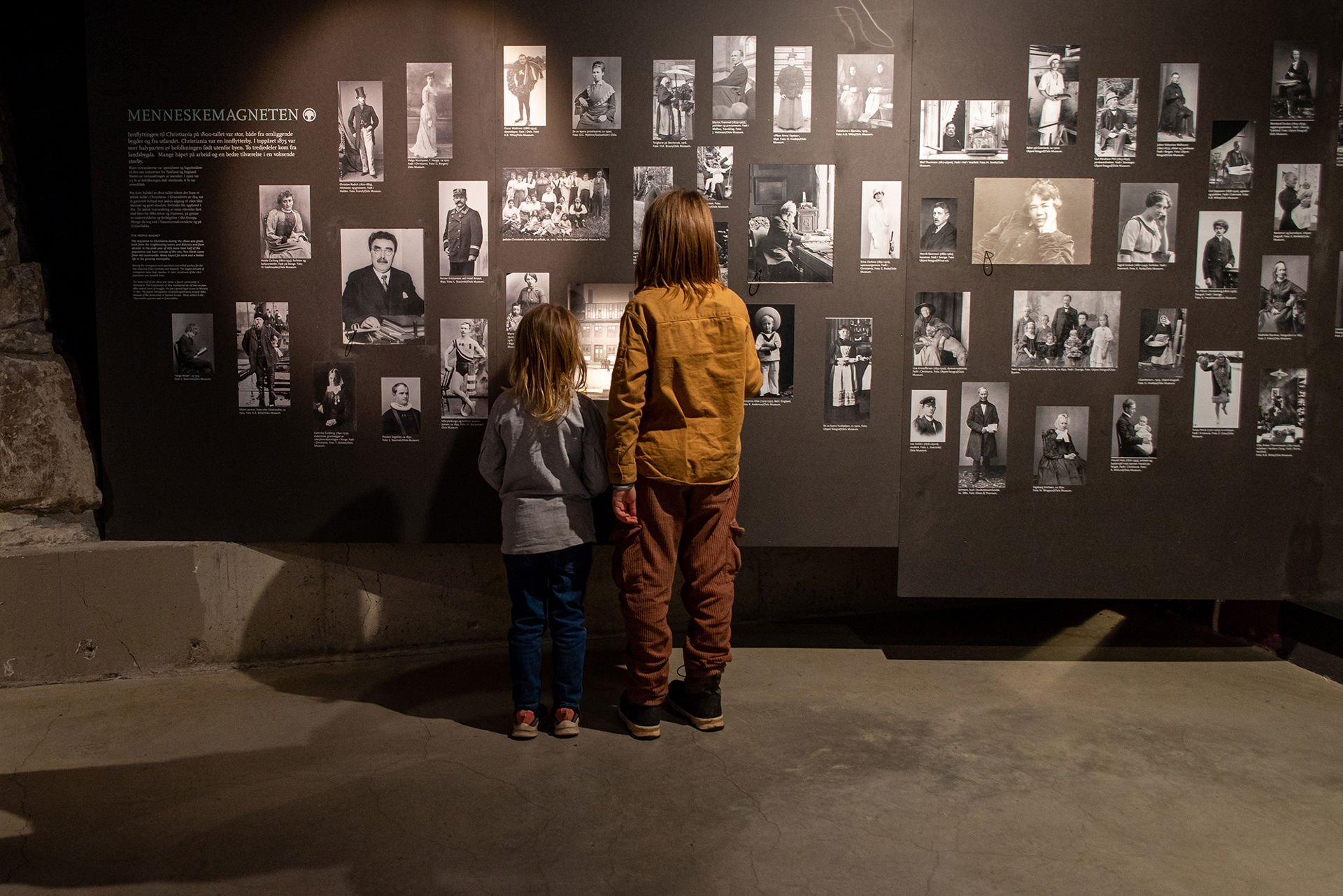 To barn ser på en vegg med mange bilder av oslopersonligheter.