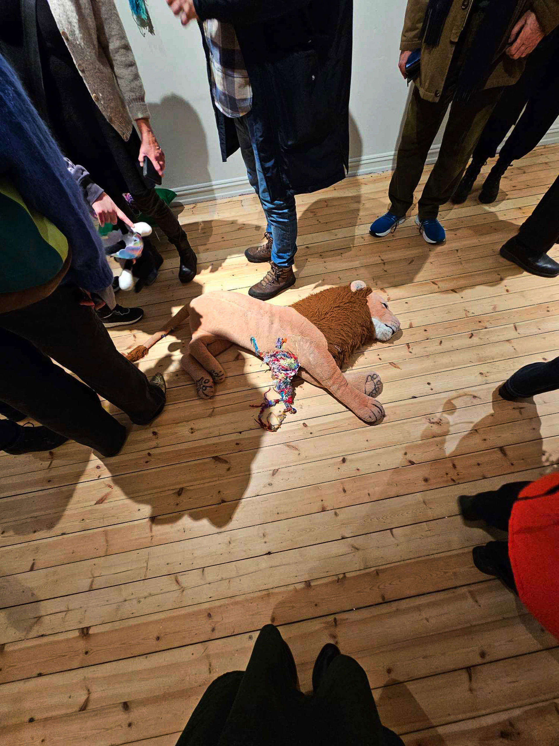 Mange mennesker i utstillingen, man ser kun ben og føtter på menneskene. En stor leketøysløve ligger på gulvet.