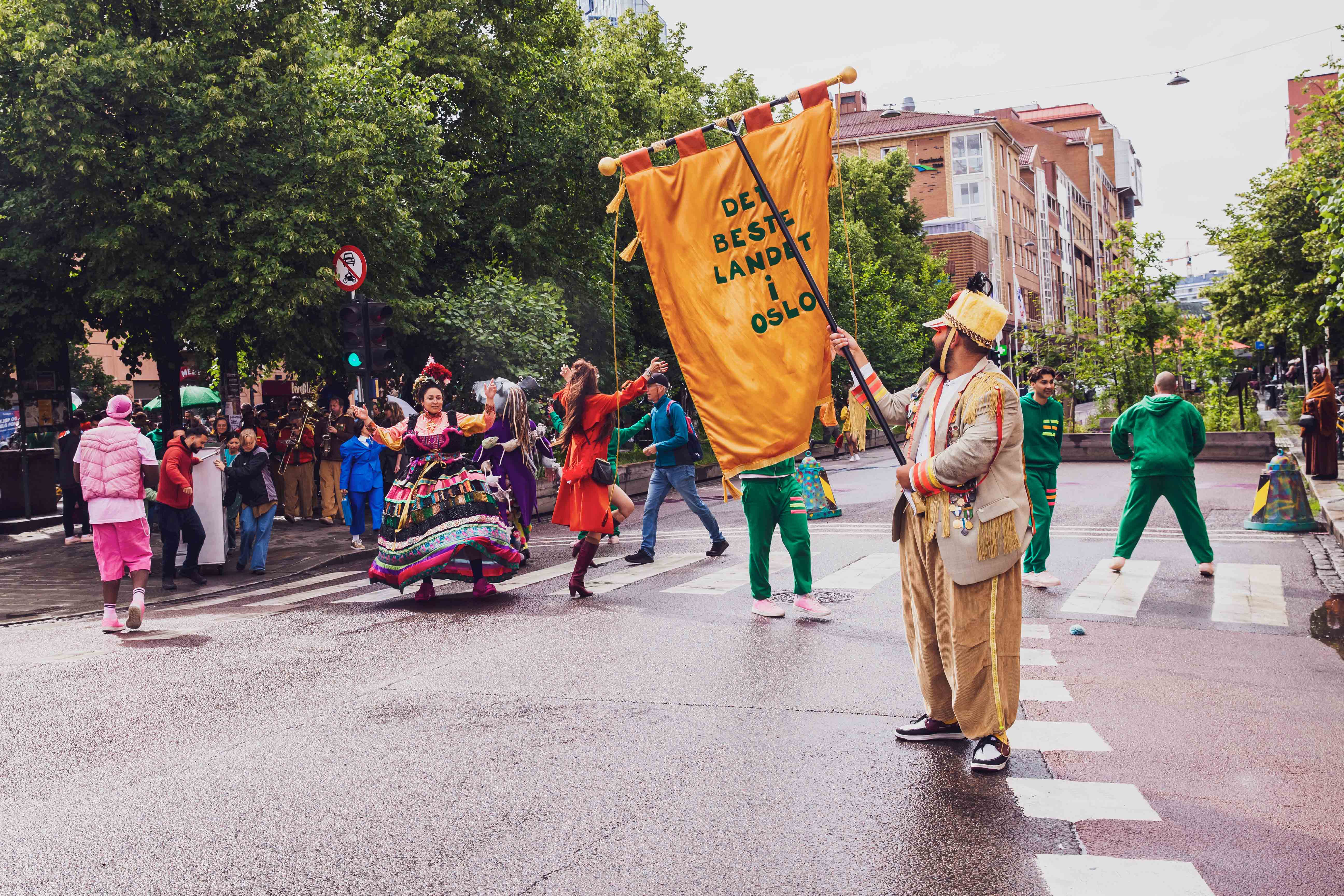 Personer kledd i fargerike klær danser over et fotgjengerfelt i en gate. En mann i gult antrekk og hatt på hodet står i forgrunnen med en gul fane der det står Det beste landet i Oslo.