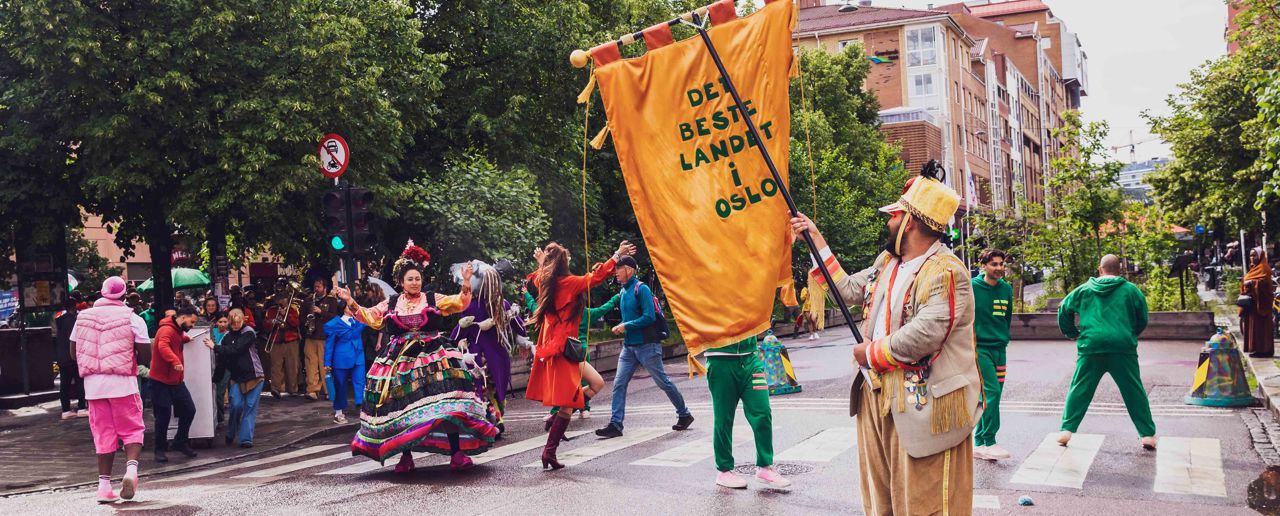 Personer kledd i fargerike klær danser over et fotgjengerfelt i en gate. En mann i gult antrekk og hatt på hodet står i forgrunnen med en gul fane der det står Det beste landet i Oslo.