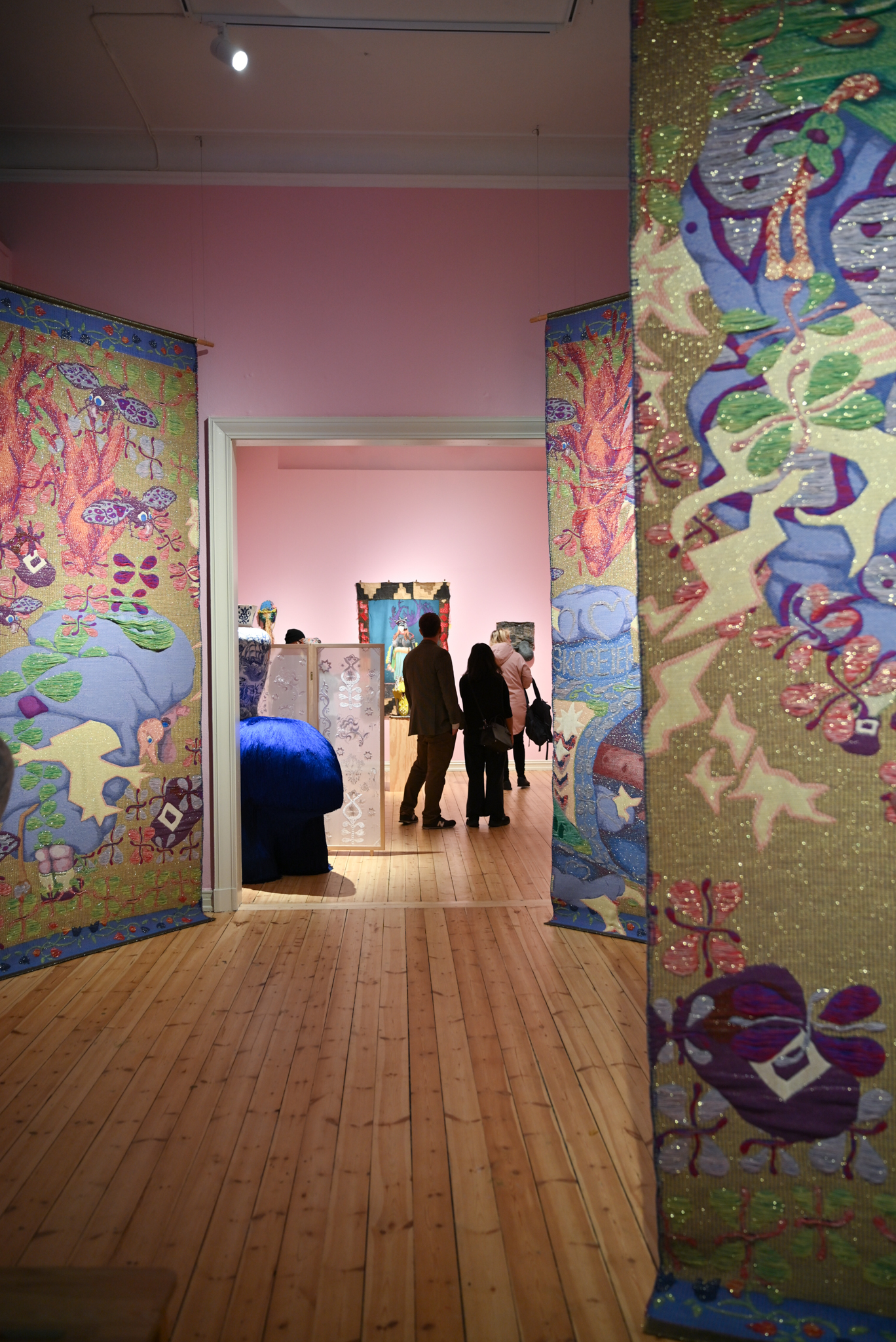 Tekstilkunst på rosa vegg bakerst, dekorert vase på blå dyreformet figur i midten, fargerik og glitrende tekstilkunst fra tak til gulv på sidene