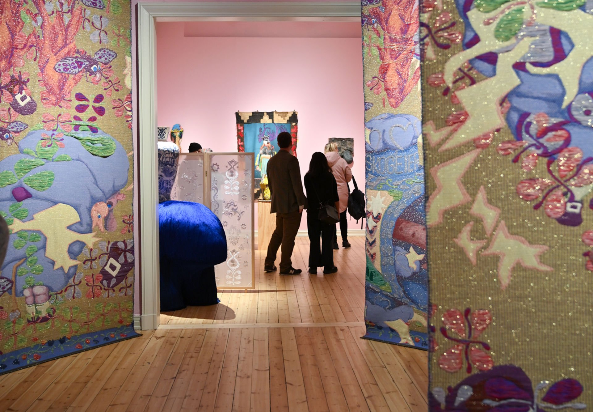 Tekstilkunst på rosa vegg bakerst, dekorert vase på blå dyreformet figur i midten, fargerik og glitrende tekstilkunst fra tak til gulv på sidene