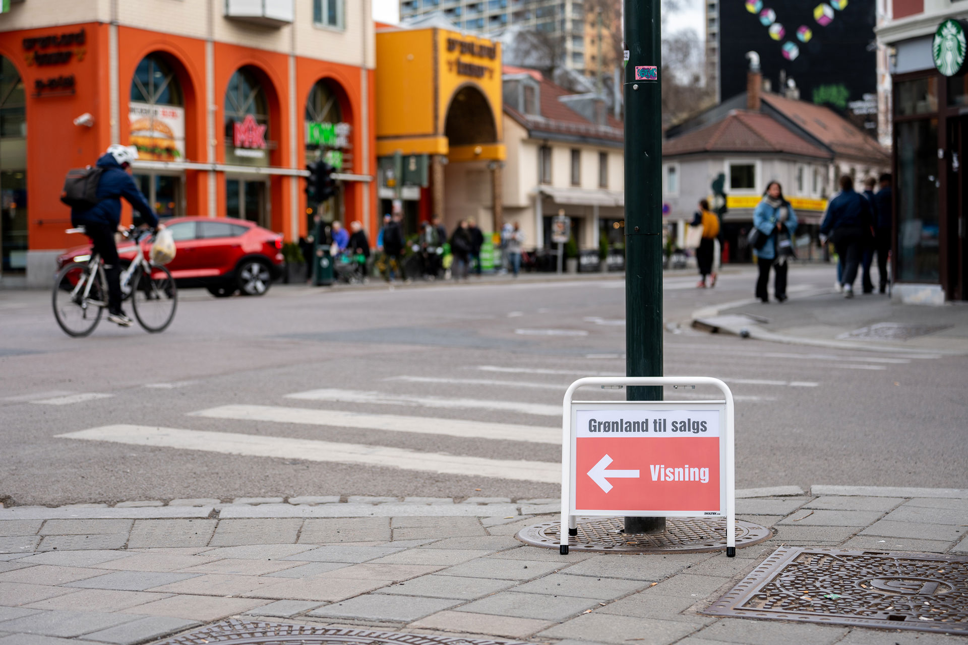 Rødt eiendomsmeglerskilt med teksten Grønland til salgs og pil til visning står på et folkerikt torg.