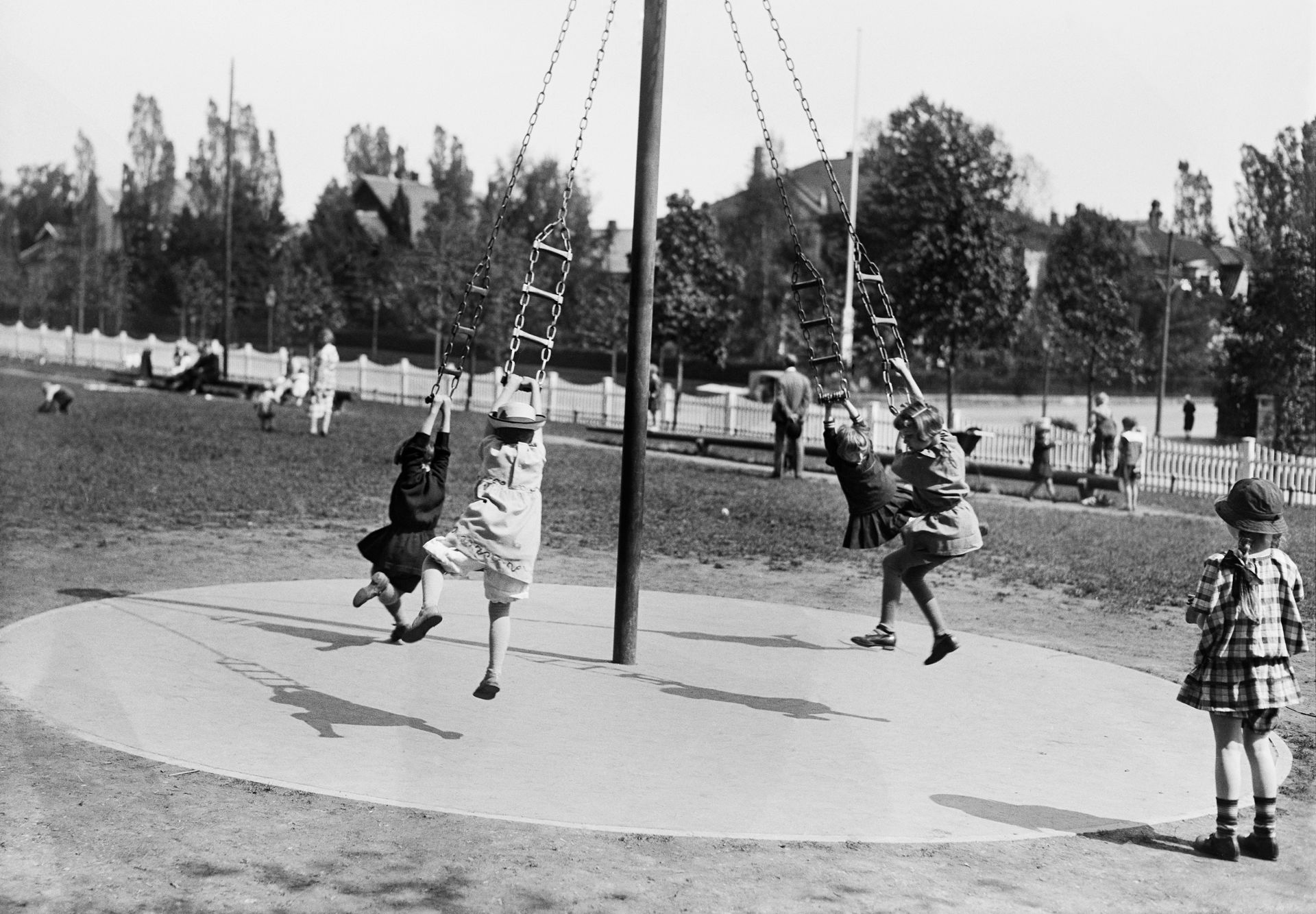 Sort-hvitt-bilde av barn i kjoler og hatter som svinger seg i et lekestativ i en park