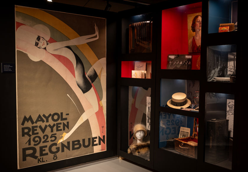 Stor plakat med illustrert kvinne og teksten Mayol-revyen 1925 Regnbuen kl. 8. Glassmontere med gjenstander til høyre.