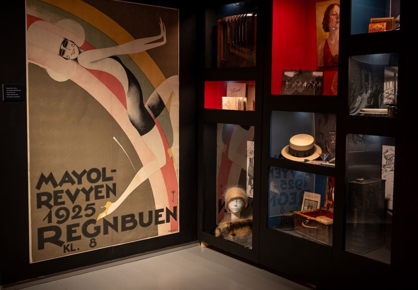 Stor plakat med illustrert kvinne og teksten Mayol-revyen 1925 Regnbuen kl. 8. Glassmontere med gjenstander til høyre.