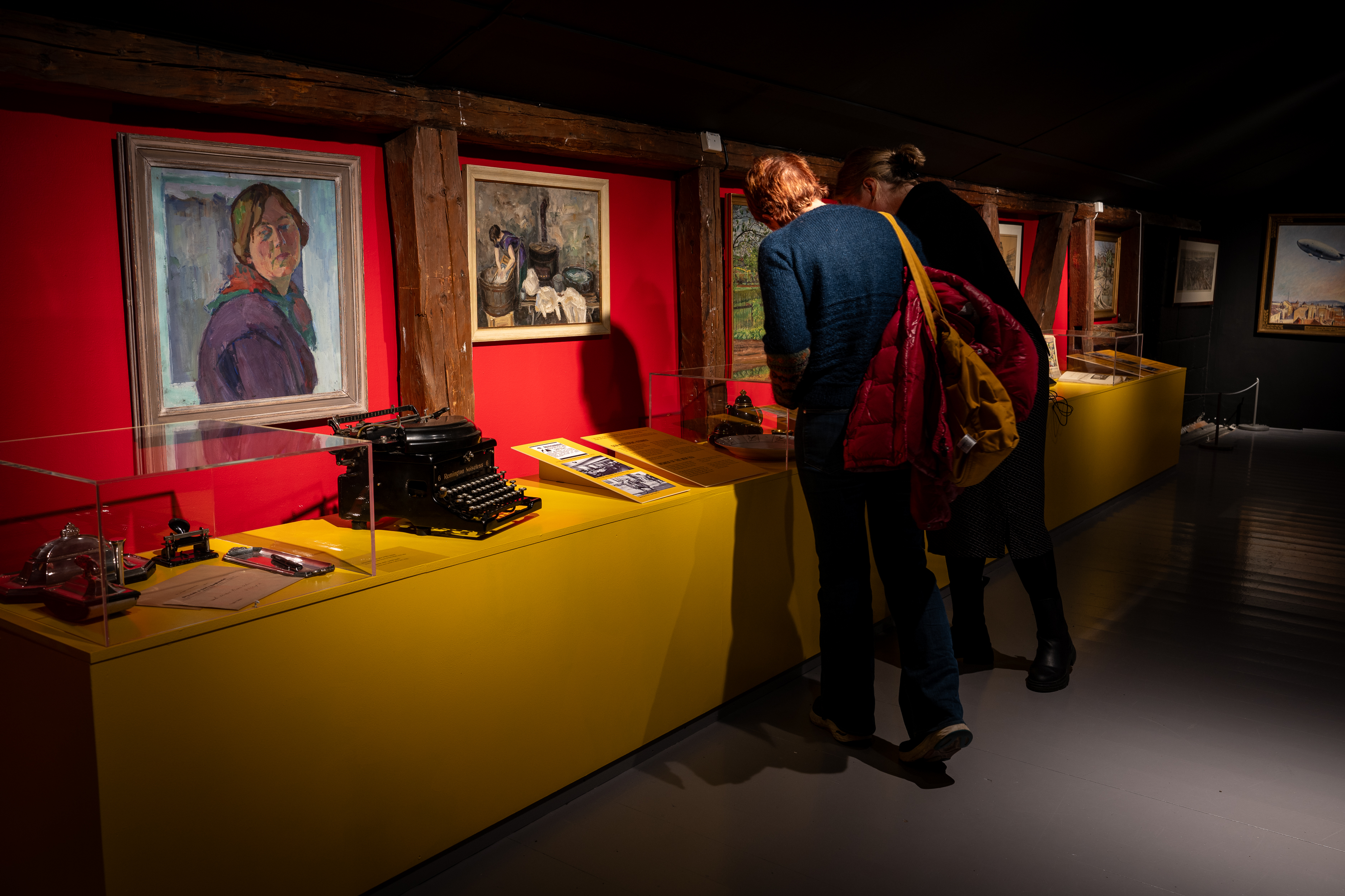 To personer står foran en rød vegg med malerier, et gult utstillingsmonter foran med gamle gjenstander. 