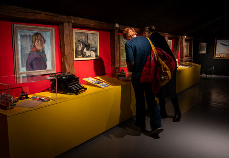 To personer står foran en rød vegg med malerier, et gult utstillingsmonter foran med gamle gjenstander.