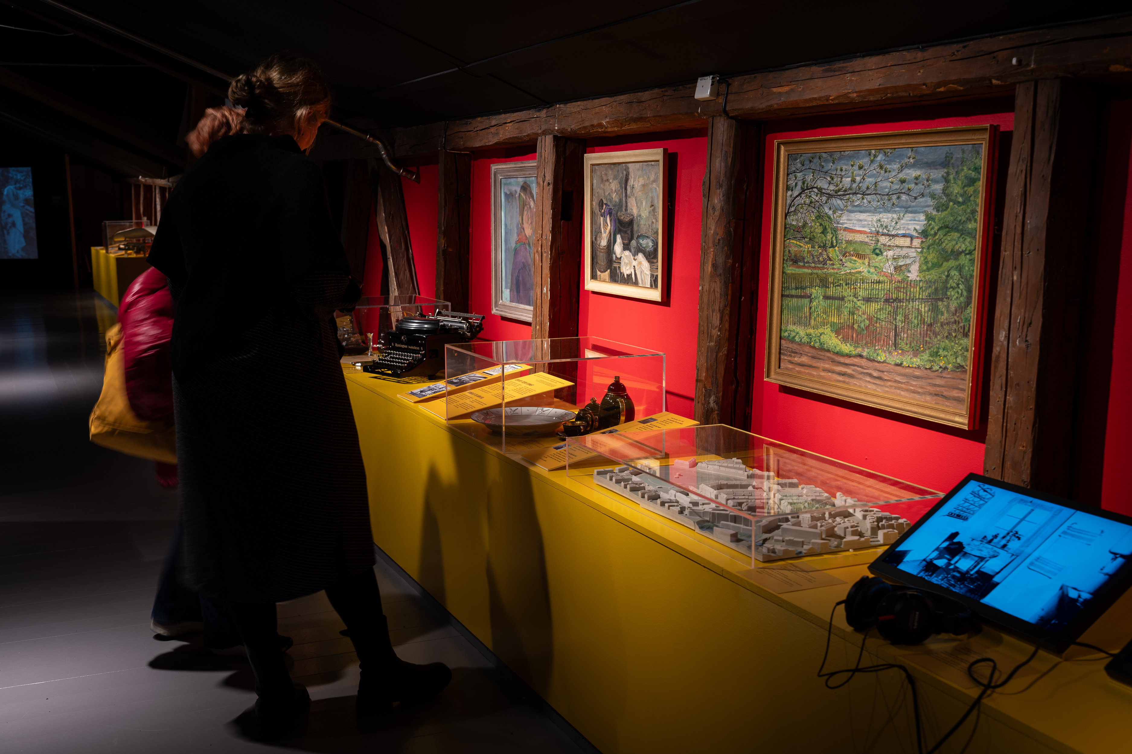 To personer med ryggen til ser ned på gjenstander i utstillingsmonter. Malerier på rød vegg foran dem