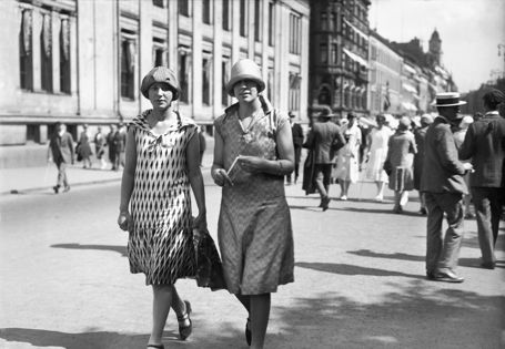 To damer kledd i 20-tallskjoler og hatter spaserer bortover Karl Johan
